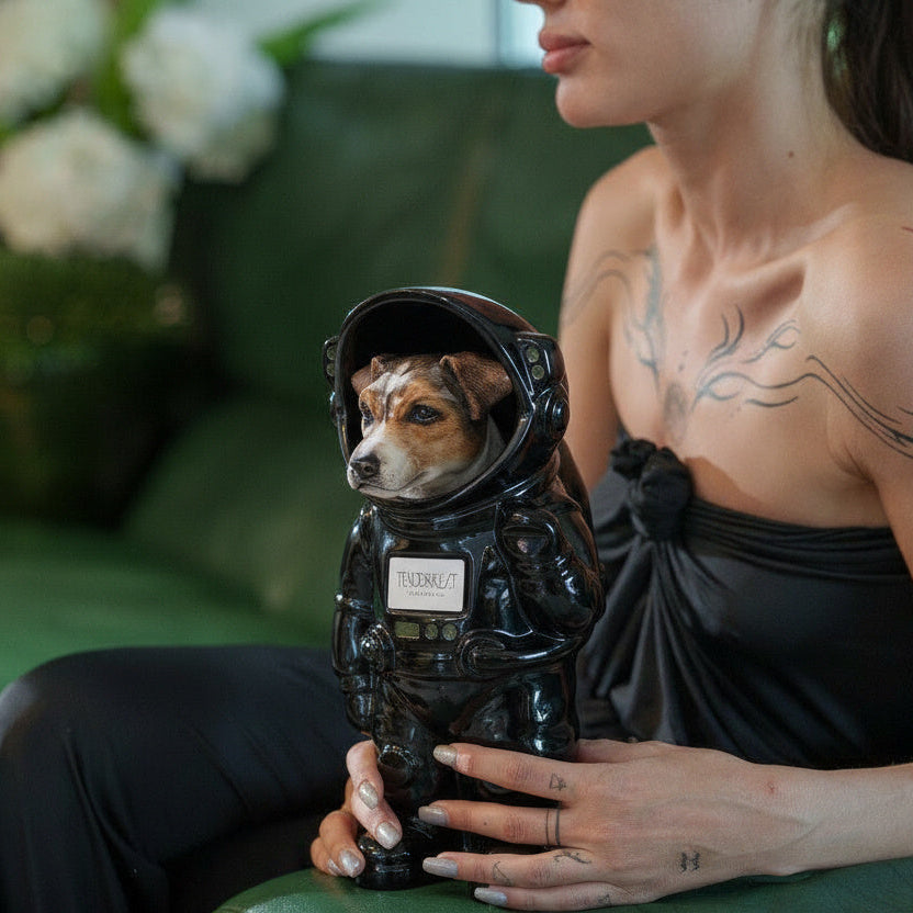 A small dog wearing a glossy black astronaut costume sits on a green leather sofa next to a woman in a black strapless dress. The dog has a name tag on its chest, and the woman has floral tattoos on her shoulder.